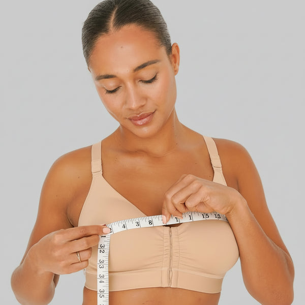 Woman measuring herself with a tape measure wearing a beige bra and panties set on a plain background