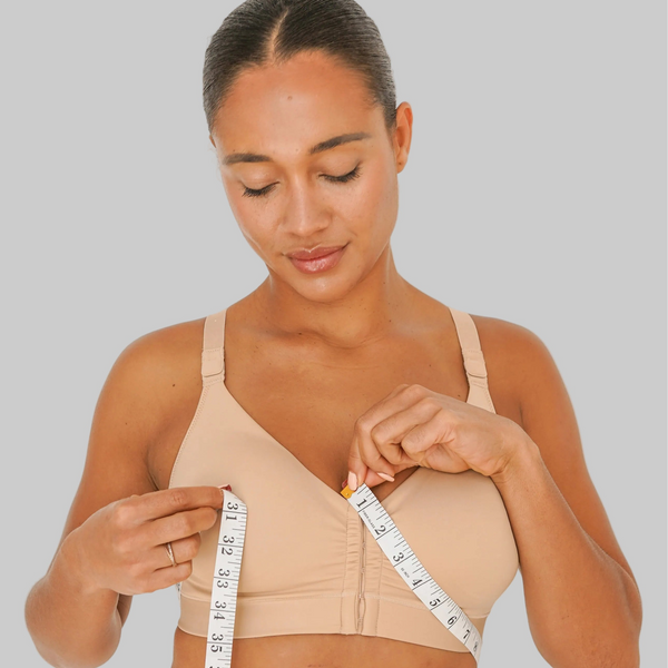 Woman measuring herself with a tape measure wearing a beige bra and panties set on a plain background